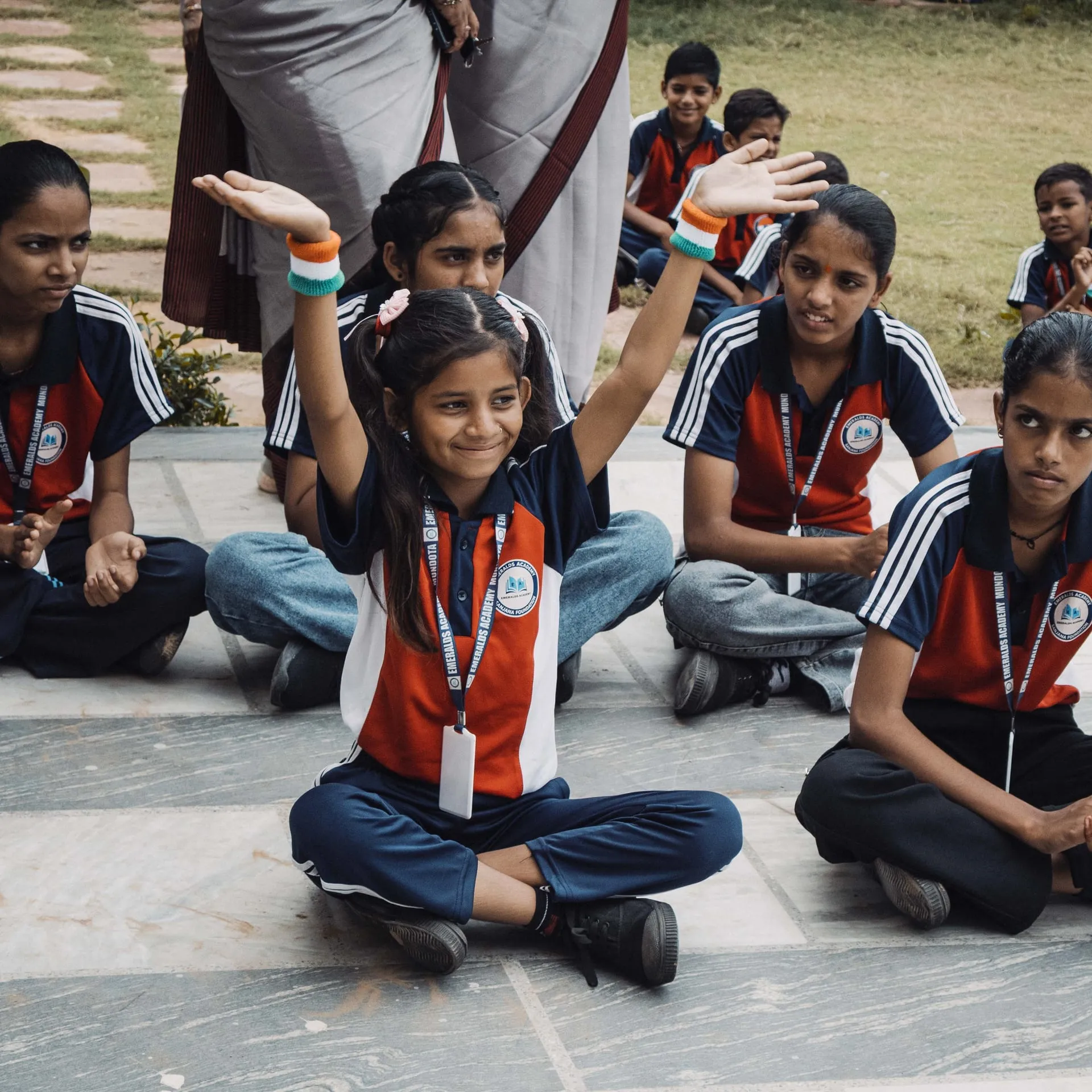 A smiling schoolgirl in uniform with both arms raised, seated among her classmates on a paved courtyard.
