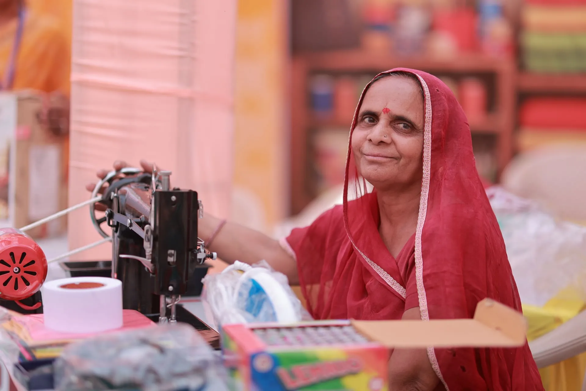 A woman in a red sari smiling beside her sewing machine at a market stall.