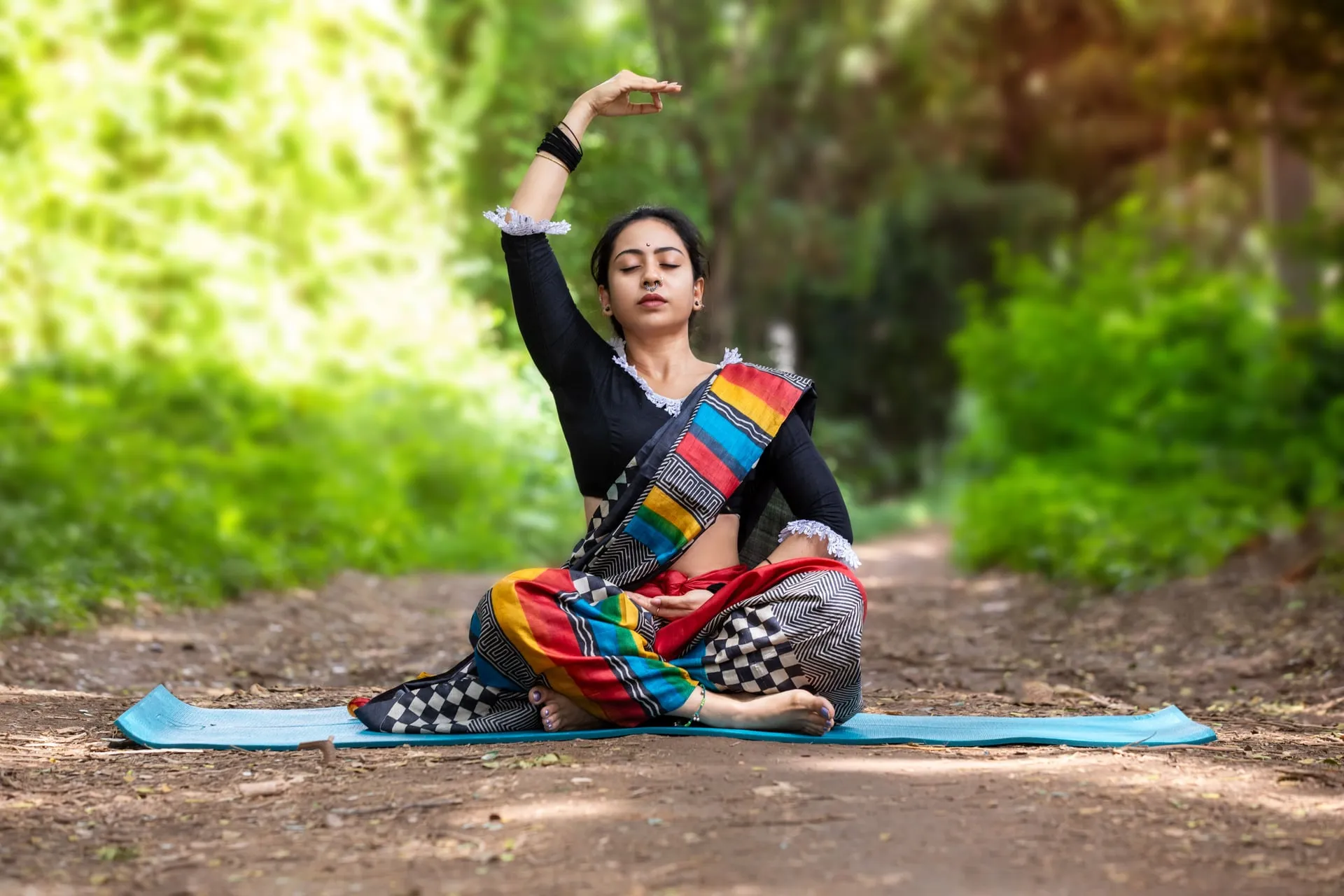 A woman in a colourful sari seated in a yoga pose on a mat outdoors, one arm raised, trees behind her.
