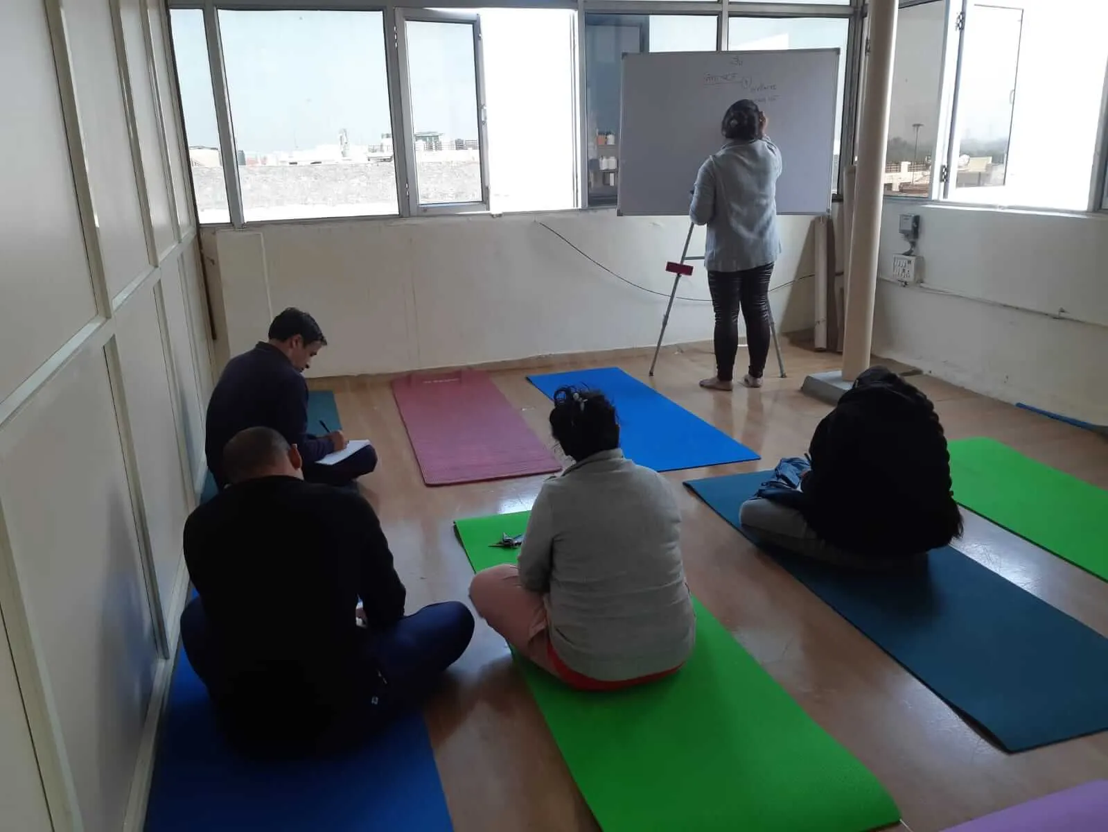 A small group seated on mats in a bright room, a teacher standing at the far wall.
